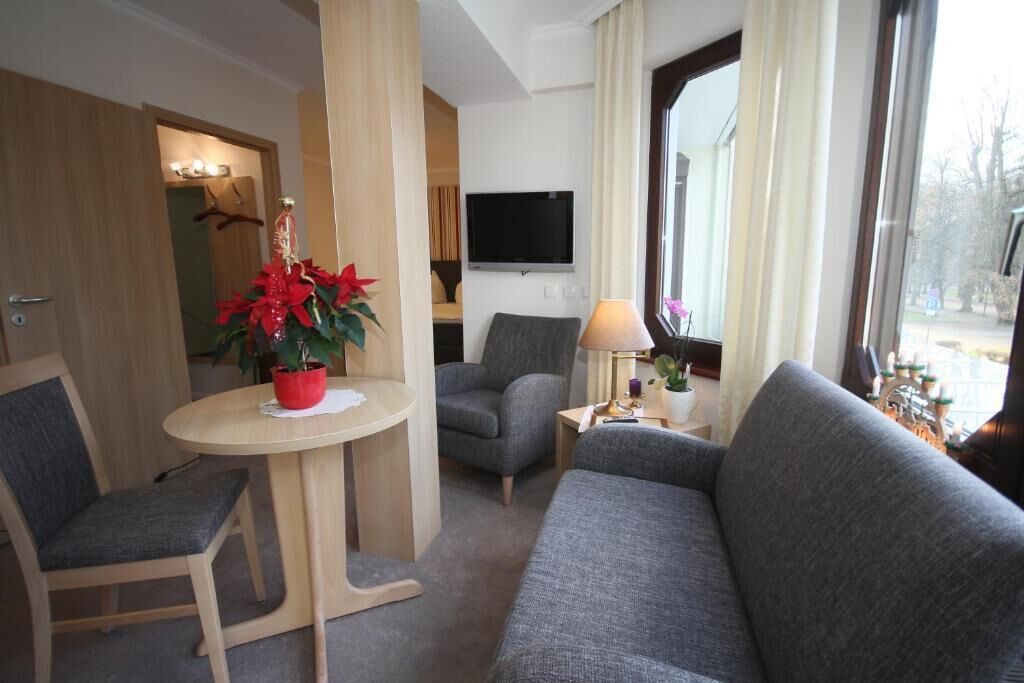 A hotel room with a sofa, a chair, a small table with a plant, a lamp, and a TV mounted on the wall.