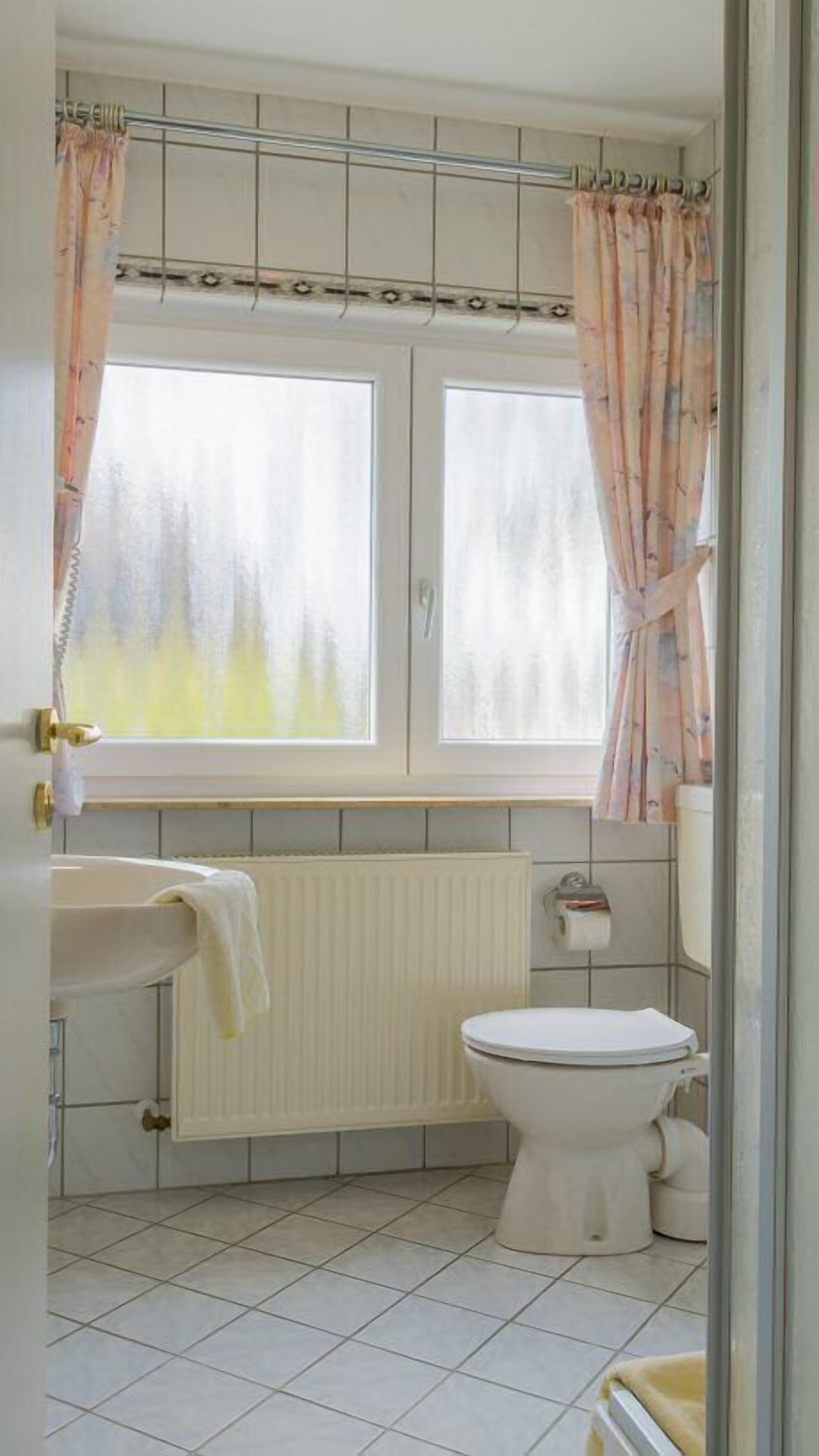 Une salle de bain comprenant des toilettes, un lavabo, une fenêtre avec des rideaux et un radiateur.