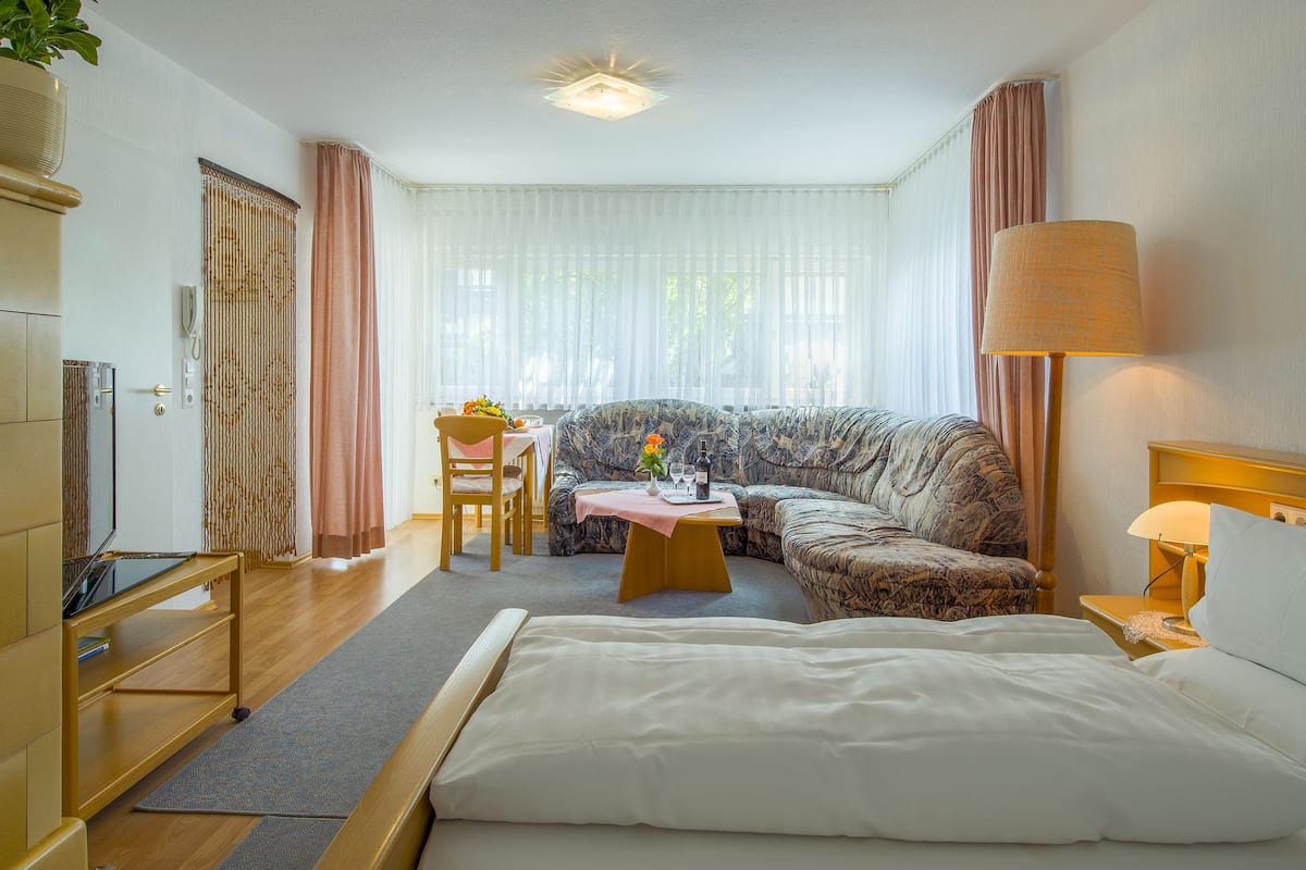 Ein Hotelzimmer mit einem Bett, einem Sofa, einem Tisch mit einer Vase, einer Lampe und einem Fernseher auf einem Standfuß.