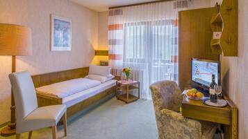 Ein gemütliches Hotelzimmer mit Bett, Sessel, kleinem Tisch mit Flasche und Gläsern, Fernseher und einem Fenster mit Vorhängen.