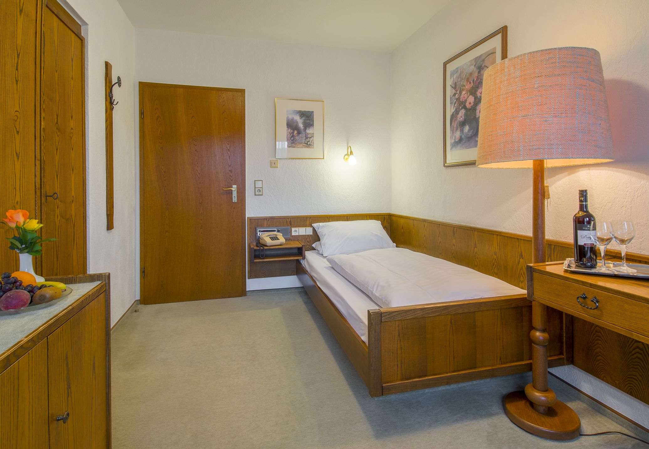 A neatly arranged bedroom with a wooden bed, a bedside table with a lamp, a nightstand with a telephone, and a wall-mounted picture.