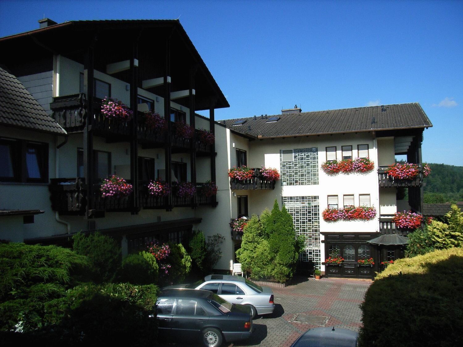 Un edificio de dos pisos con balcones decorados con macetas de flores, un camino de entrada pavimentado y coches estacionados.