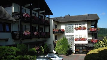Un edificio de dos pisos con balcones decorados con macetas de flores, un camino de entrada pavimentado y coches estacionados.