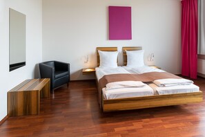 A modern hotel room with a wooden bed, a black chair, a nightstand, and a pink wall art piece. - AMH Airport-Messe-Hotel (Filderstadt)