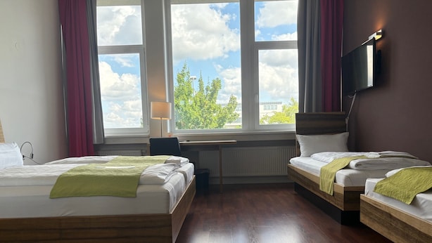 A hotel room with two beds, a desk, a chair, a TV mounted on the wall, and a window with a view of trees and buildings. - AMH Airport-Messe-Hotel (Filderstadt)