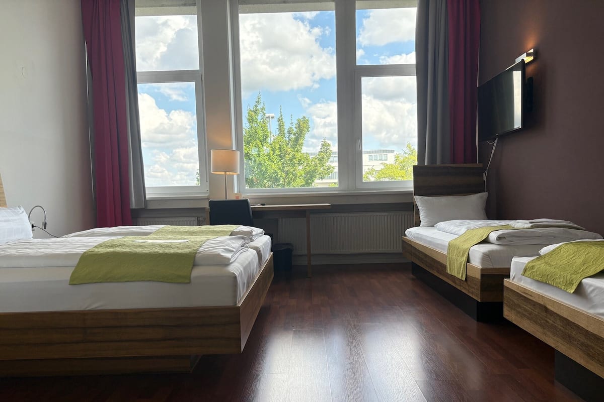 A hotel room with two beds, a desk, a chair, a TV mounted on the wall, and a window with a view of trees and buildings.