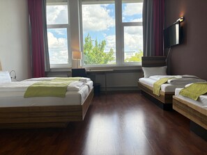 A hotel room with two beds, a desk, a chair, a TV mounted on the wall, and a window with a view of trees and buildings. - AMH Airport-Messe-Hotel (Filderstadt)