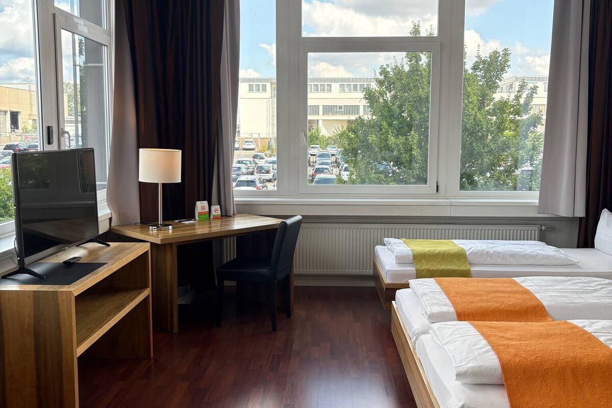 A hotel room with a wooden desk, a TV, a bed with orange and white bedding, and large windows offering a view of the city.