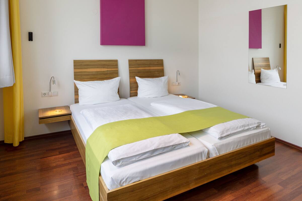 A double bed with a wooden frame, two bedside tables with lamps, a mirror on the wall, and a yellow curtain.