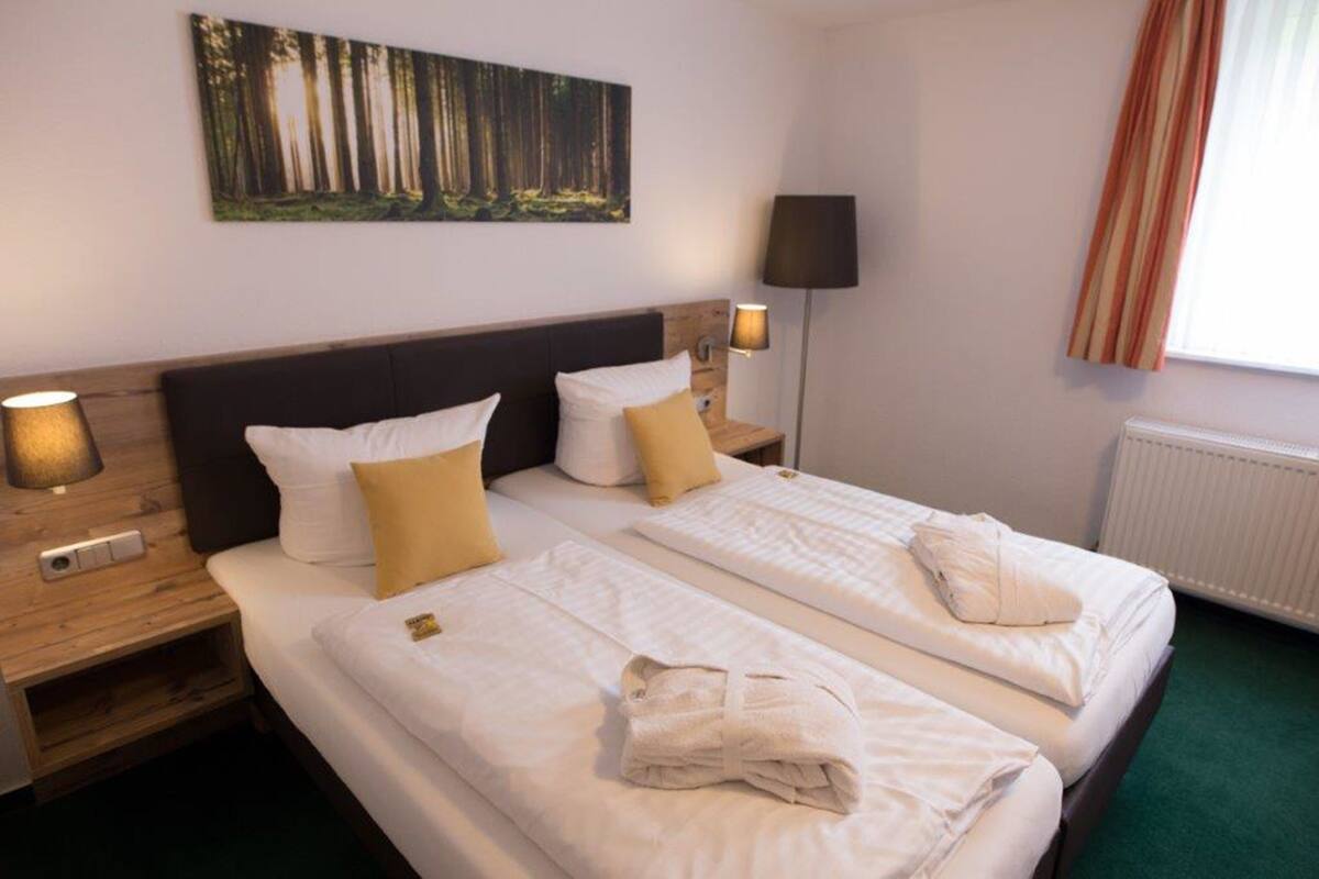 Een hotelkamer met twee bedden, een nachtkastje met een lamp, een schilderij aan de muur en een raam met gordijnen.