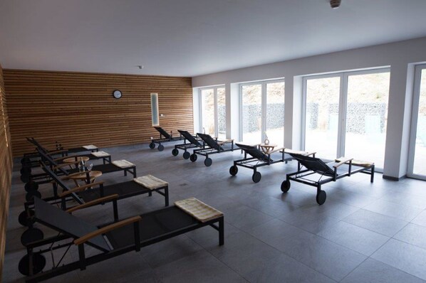 A spacious indoor area with multiple sun loungers and tables, large windows allowing natural light, and a wooden panel wall. - BSW - Erlebnishotel Festenburg (Clausthal-Zellerfeld)