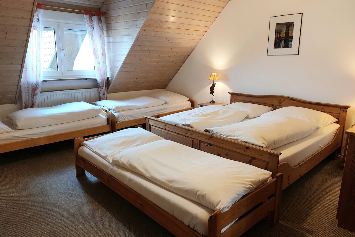 Ein Zimmer mit drei Holzbett, einem Heizkörper, einem Fenster mit Vorhängen, einer Lampe und einem gerahmten Bild an der Wand.