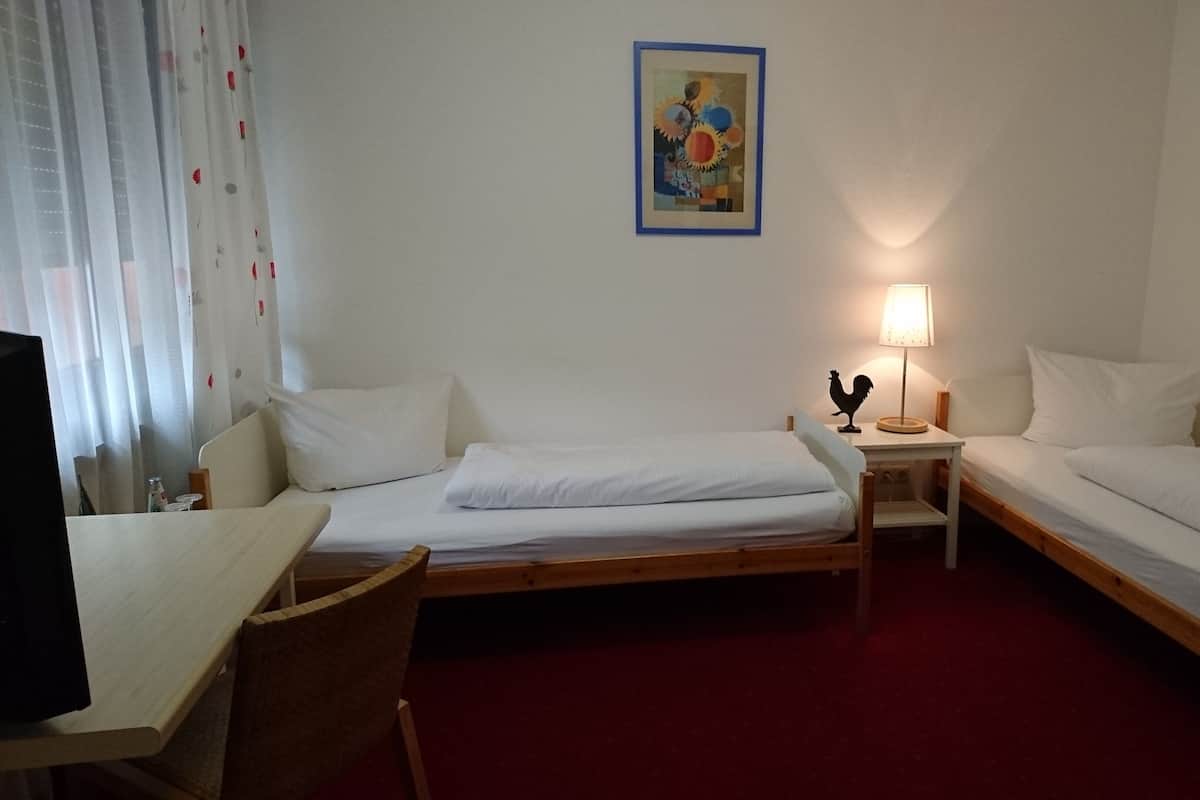 Ein Hotelzimmer mit zwei Einzelbetten, einem Schreibtisch, einem Stuhl, einer Lampe und zwei gerahmten Kunstwerken an den Wänden.