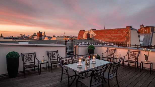 A rooftop terrace with a table set for two, surrounded by chairs, and a view of the city skyline at sunset. - Palacina Berlin Apartments (Berlin)