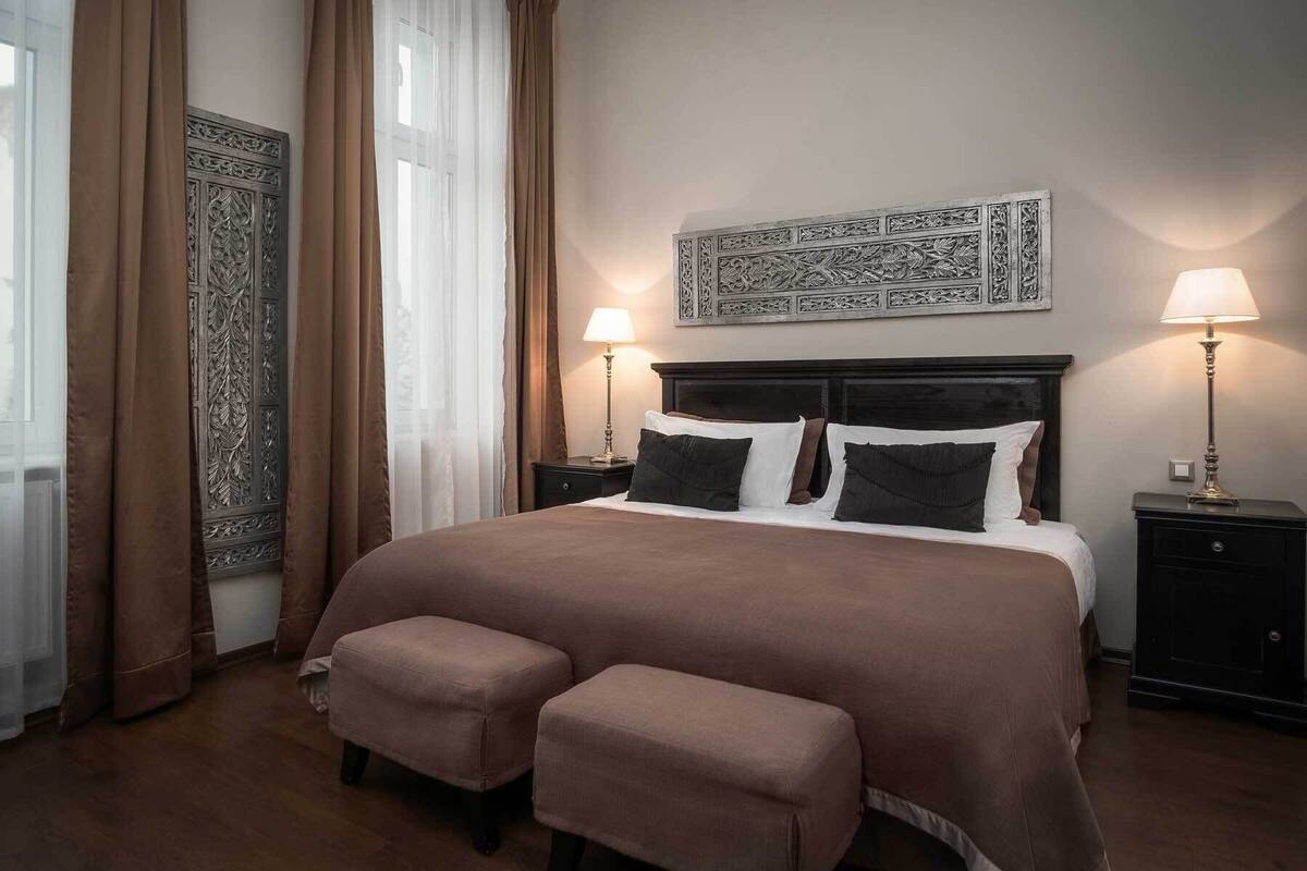 Ein Hotelzimmer mit einem großen Bett, zwei Nachttischen inklusive Lampen, einer verzierten Kopfteil, einem großen Fenster mit Vorhängen und einer kleinen Bank.