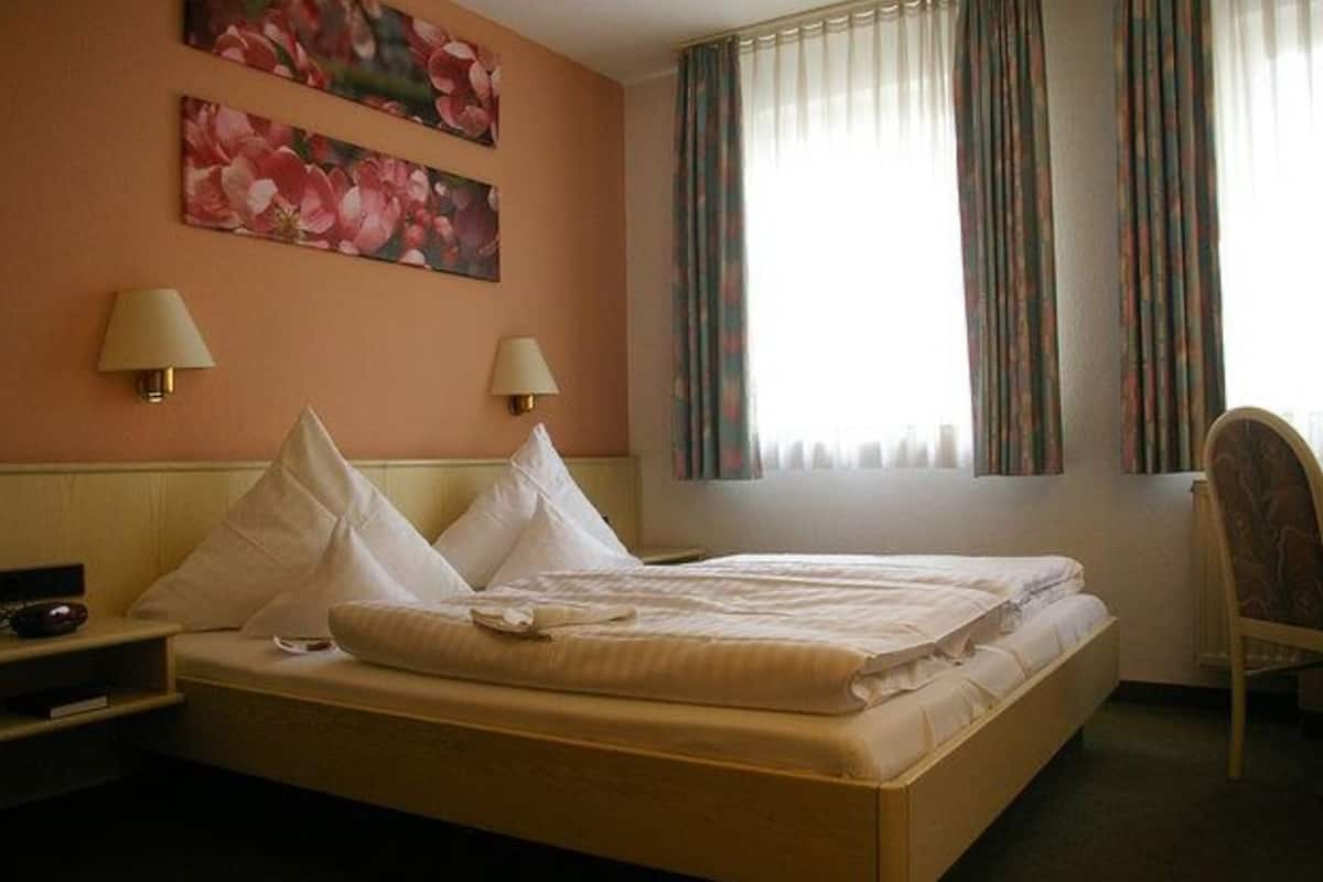 Una habitación de hotel con una cama, dos lámparas de noche, una silla y un ventanal con cortinas.