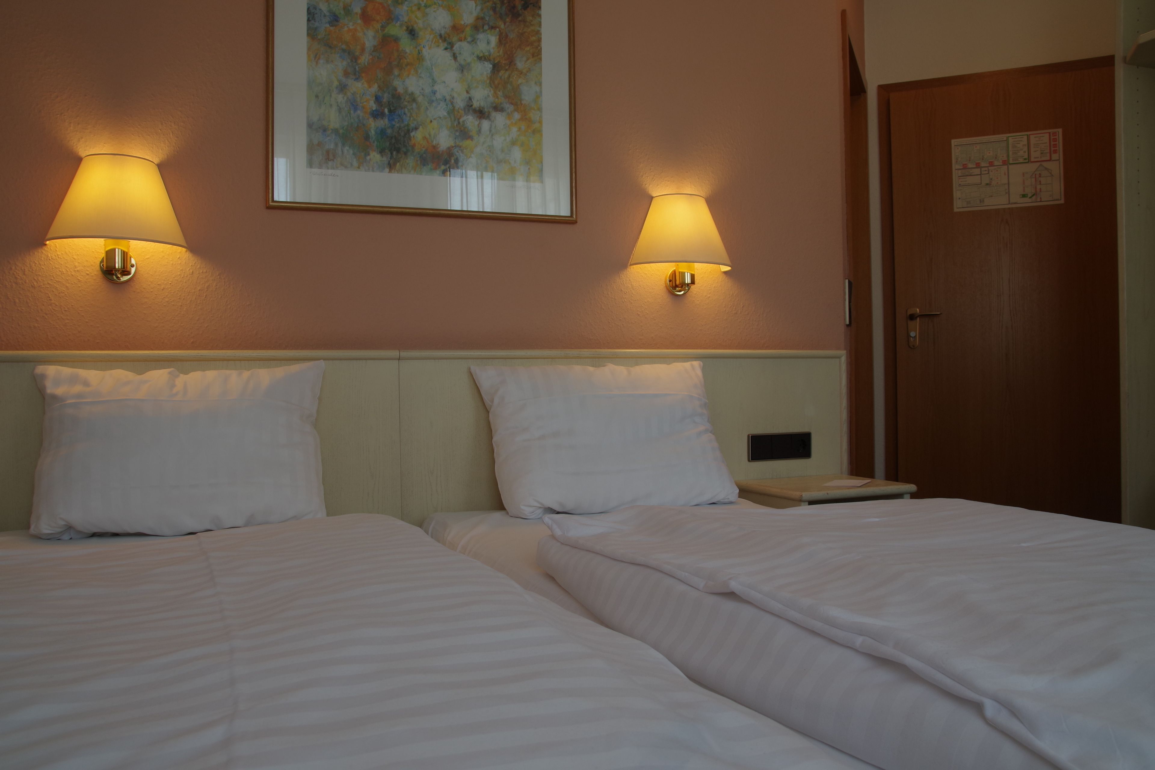 Ein Hotelzimmer mit zwei Betten, jeweils mit weißer Bettwäsche und Kissen. An jeder Seite der Betten befinden sich Wandlampen.