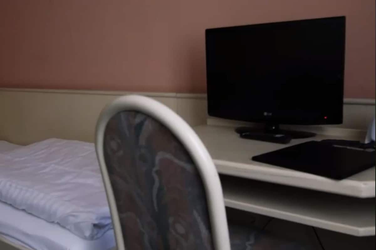 Una cama individual con ropa de cama blanca, un televisor sobre un escritorio y una silla con respaldo estampado.