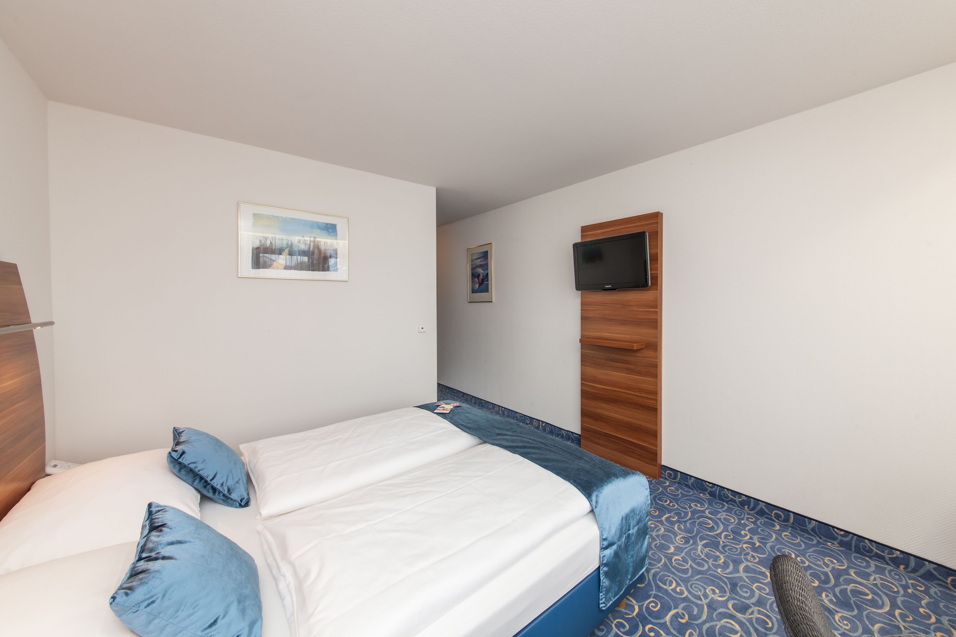 Habitación de hotel con una cama, dos almohadas, un televisor montado en la pared y una alfombra estampada.