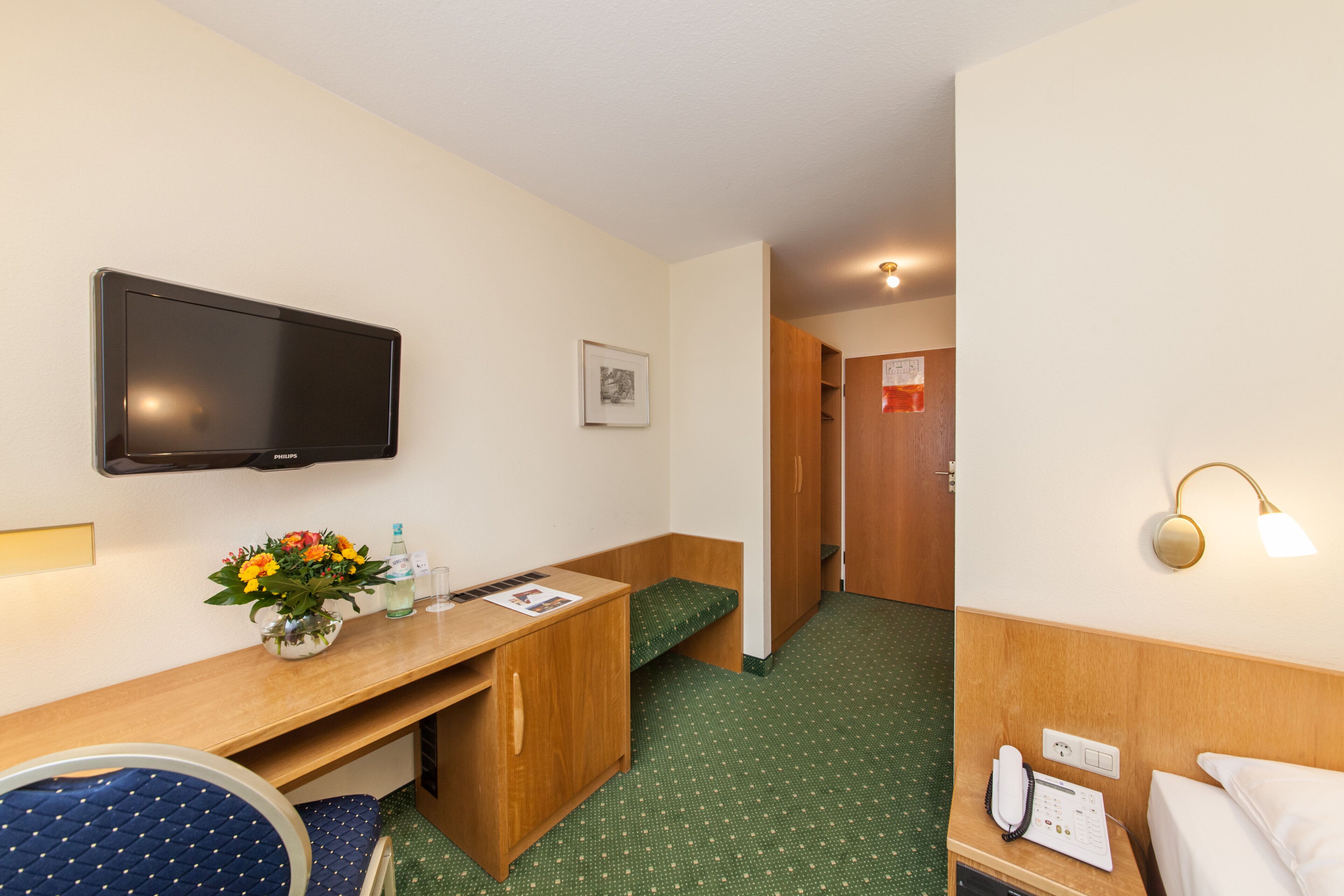 A hotel room with a bed, a desk with a chair, a television, a bedside table with a lamp, a small window, and a door leading to the hallway.
