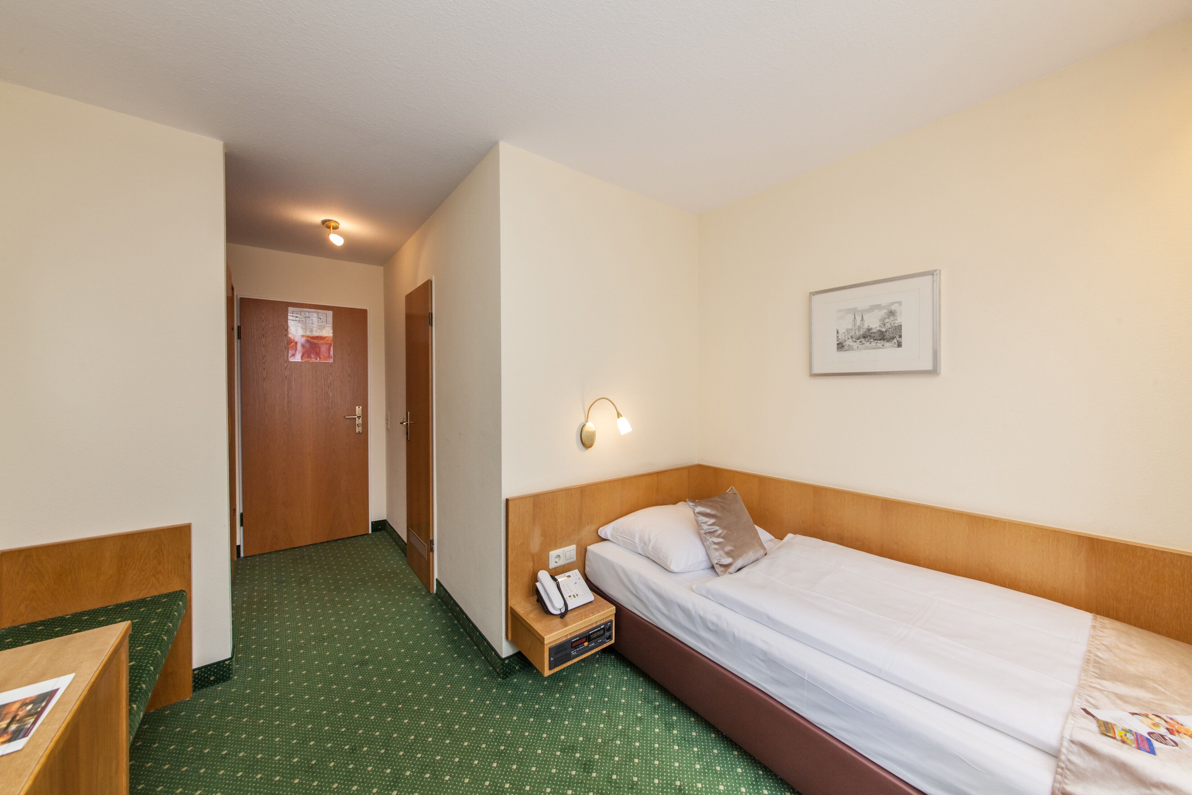 Una camera d'albergo con un letto singolo, un comodino con il telefono, una lampada a parete e una porta che dà su un'altra stanza.