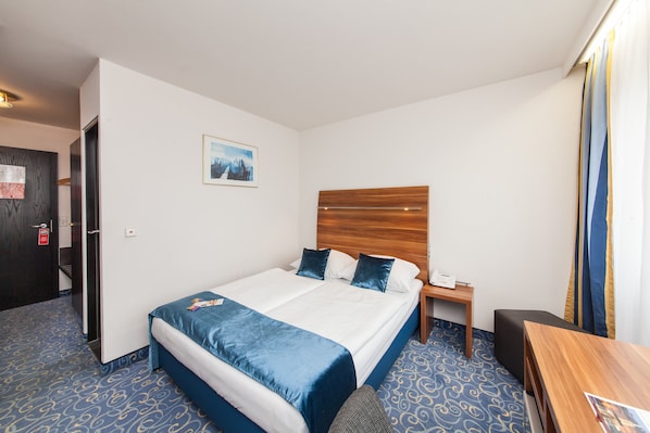 Una habitación de hotel con una cama grande, cabecera de madera, mesita de noche, escritorio y alfombra estampada.