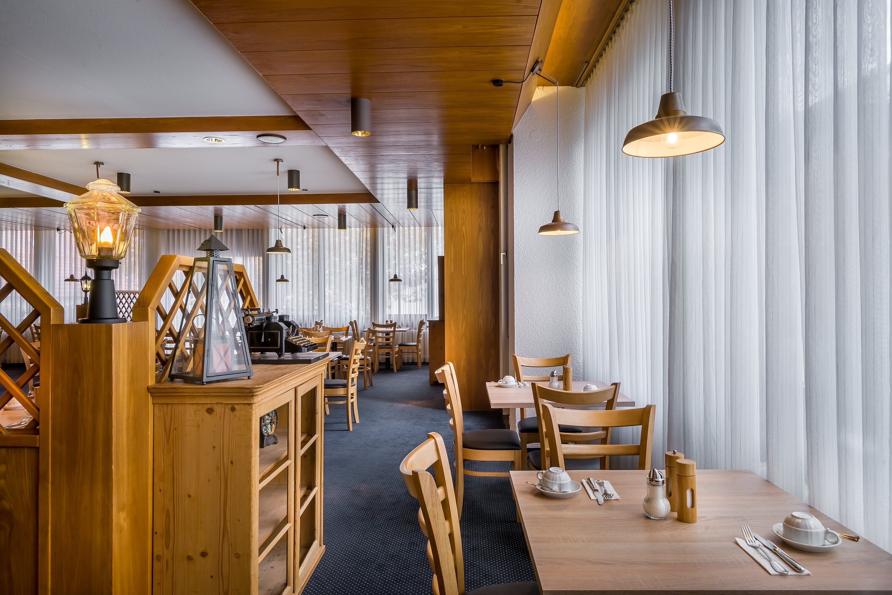 A restaurant with wooden furniture, pendant lights, and a view through large windows.