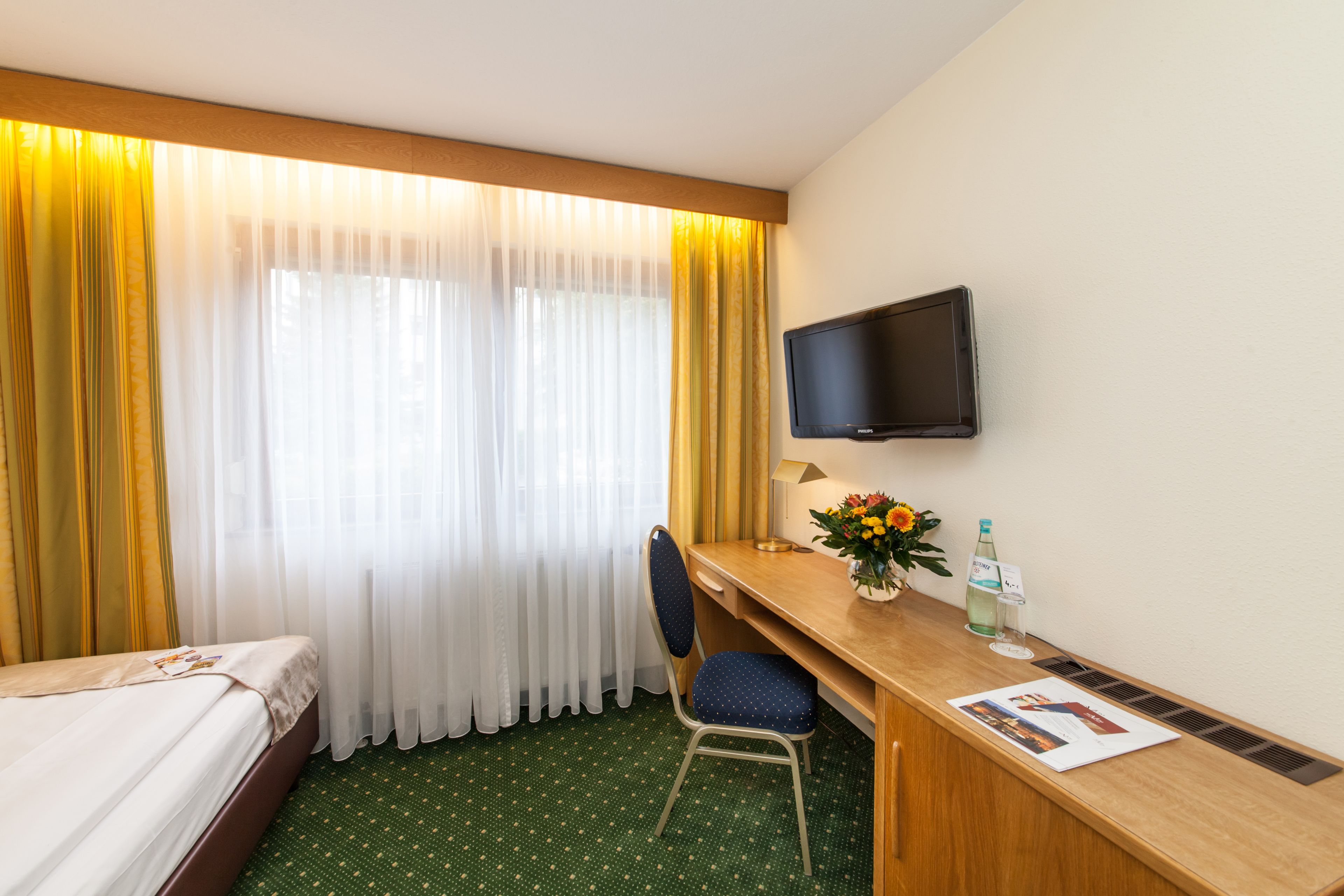 A hotel room with a bed, a desk with a chair, a television, and a window with curtains.
