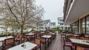 An outdoor dining area with wooden tables and chairs, surrounded by a wooden deck, overlooking a residential area with modern buildings. - Hotel Strohgäu (Korntal-Munchigan)