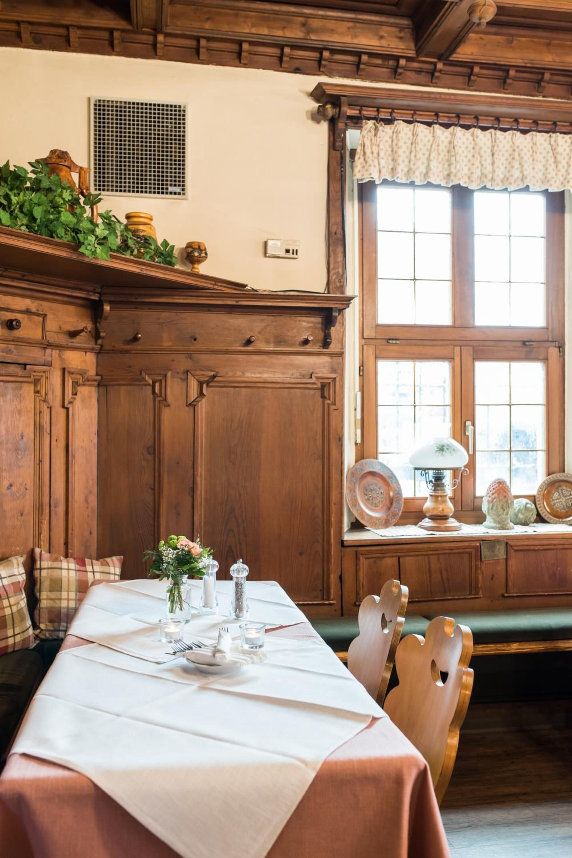 Een eetruimte met een houten tafel gedekt voor twee personen, een raam met kantgordijnen en een houten bank met een kussen.