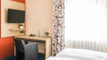Ein Hotelzimmer mit Bett, Schreibtisch mit Flachbildfernseher, Sessel, kleinem Tischchen mit Telefon, Flasche und Blumen in einer Vase.