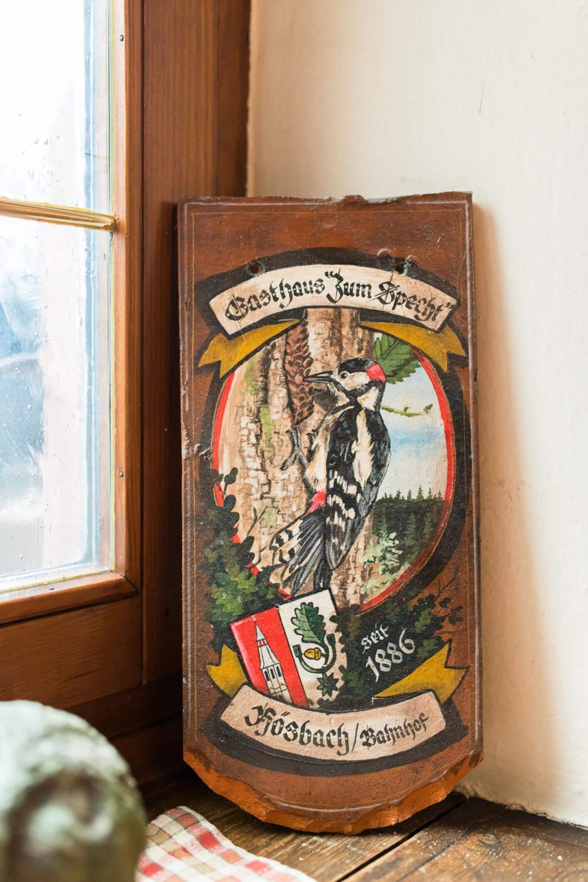 Un letrero de madera con una ilustración de un pájaro y texto, colocado sobre el alféizar de una ventana.