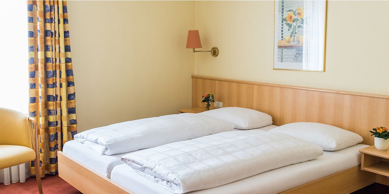 A double bed with white bedding, a wooden headboard, a nightstand with a lamp, a chair, and a framed picture on the wall.