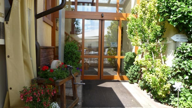 A building entrance with a glass door, potted plants, and a sign indicating WC locations. - Hotel Zum Specht (Hoesbach)