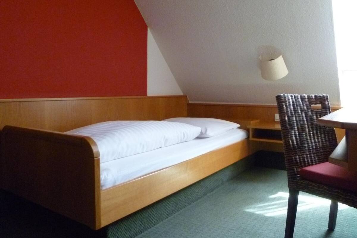 Un letto singolo con struttura in legno e biancheria bianca, una sedia in vimini con cuscino rosso e un tavolino.