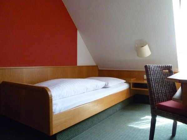 A single bed with a wooden frame and white bedding, a wicker chair with a red cushion, and a small table. - Hotel Zum Specht (Hoesbach)
