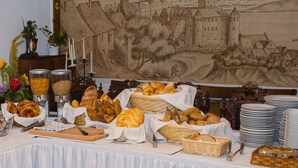 Een buffettafel met verschillende broden en gebak, een groot wandtapijt waarop een kasteel is afgebeeld en een witte tafelkleed.