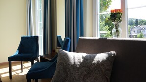 Een kamer met een bank, twee blauwe fauteuils, een kussen met een patroon, een vaas met bloemen en uitzicht op groen en gebouwen.