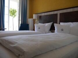 Quarto de hotel com duas camas, uma mesa de cabeceira, uma luminária e vista para uma árvore pela janela.