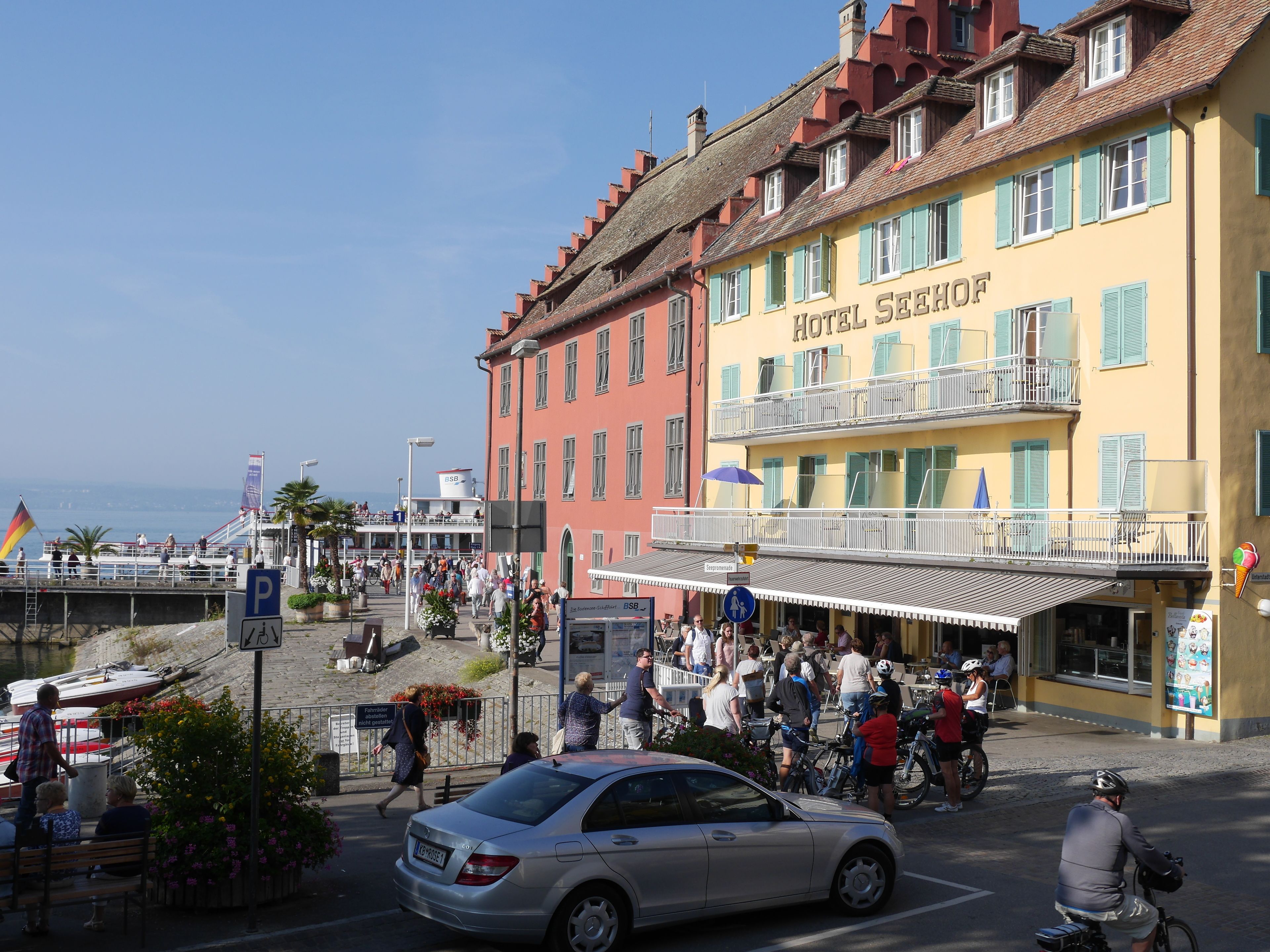 Eine belebte Straßenszene mit einem Hotel namens "HOTEL SEEHOF", das Balkone, Außenbereich und Meerblick bietet.