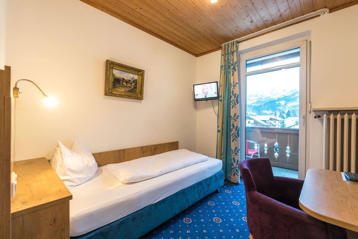 A room with a single bed, a wooden headboard, a small table, a chair, a TV, and a window with a view of mountains.
