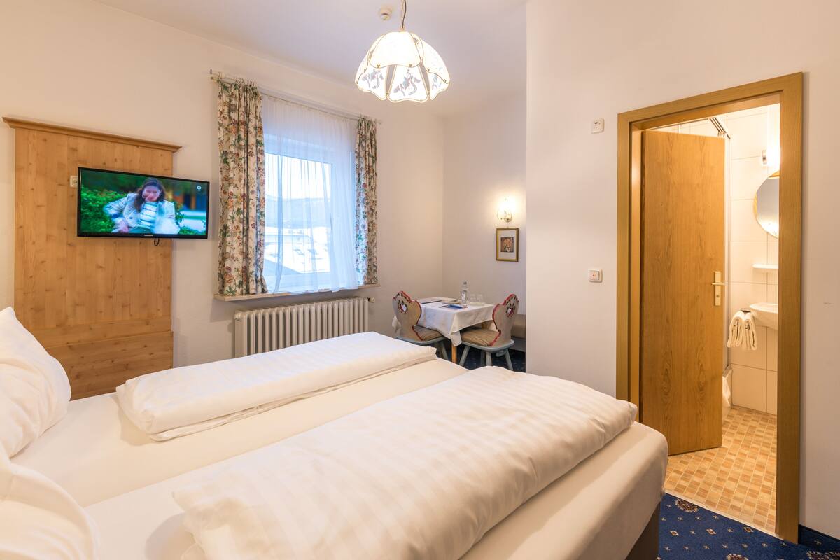 A hotel room with two beds, a television, a window with curtains, a small table with chairs, and a bathroom visible through an open door.