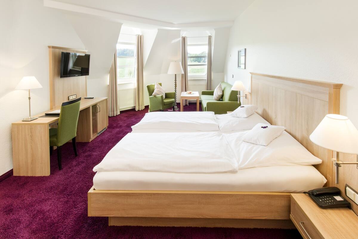 A hotel room with a large bed, two green armchairs, a desk with a computer, a TV, and a window with a view of the outdoors.