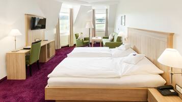 Ein Hotelzimmer mit einem großen Bett, zwei grünen Sesseln, einem Schreibtisch mit Computer, einem Fernseher und einem Fenster mit Blick ins Freie.