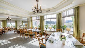 A spacious dining area with multiple tables set for a meal, large windows with curtains, and a chandelier.