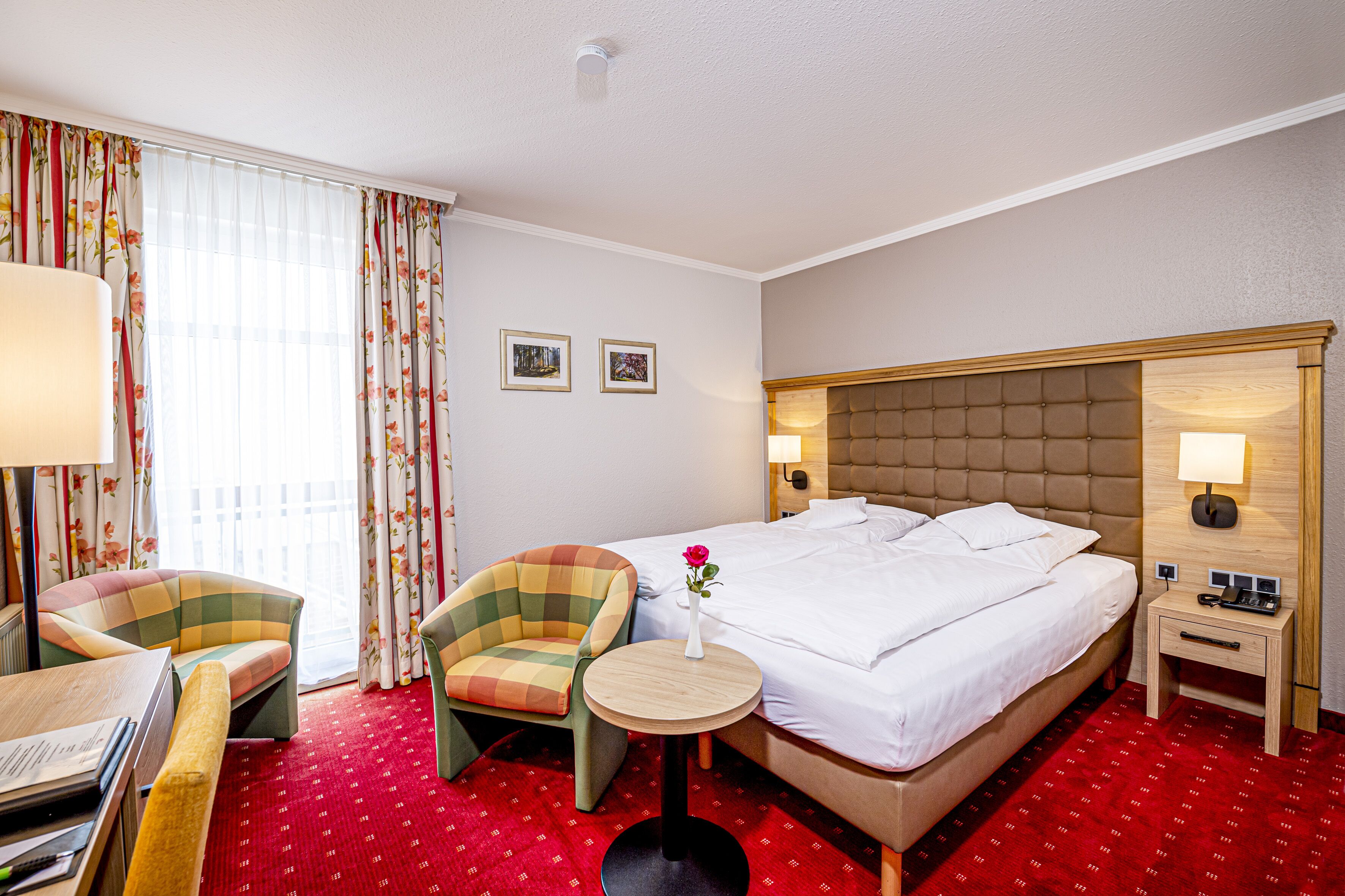 Une chambre d’hôtel avec un lit, une chaise, une petite table, une lampe et une fenêtre avec des rideaux.