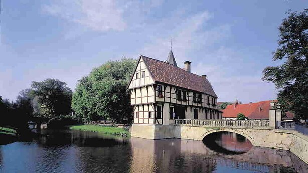 Une maison traditionnelle à colombages et au toit de chaume, située au bord d’une rivière paisible, qu’enjambe un pont de pierre.