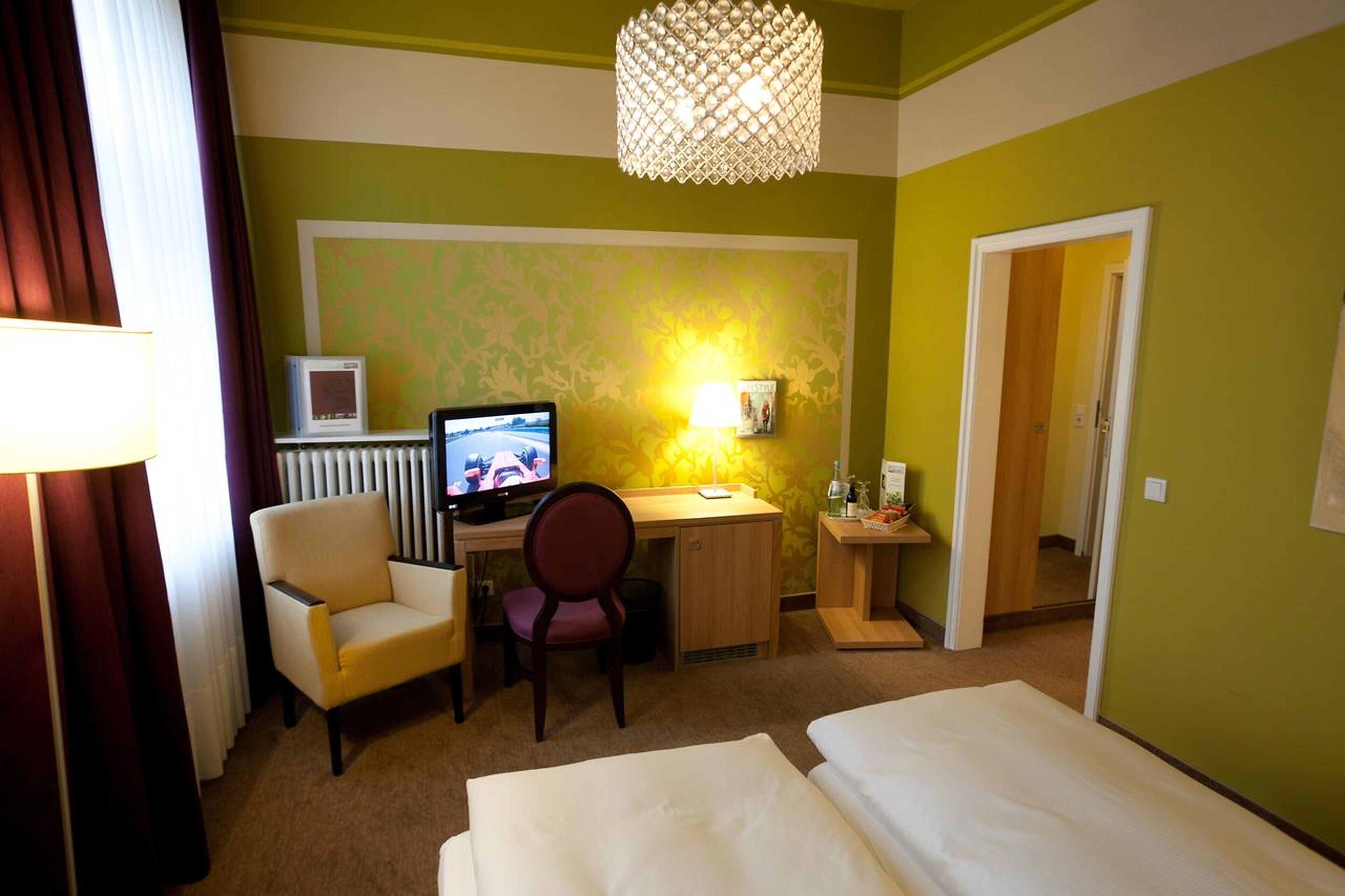 Ein Hotelzimmer mit einem Bett, einem Schreibtisch mit Computer, einem Sessel und einem Fenster mit Vorhängen.