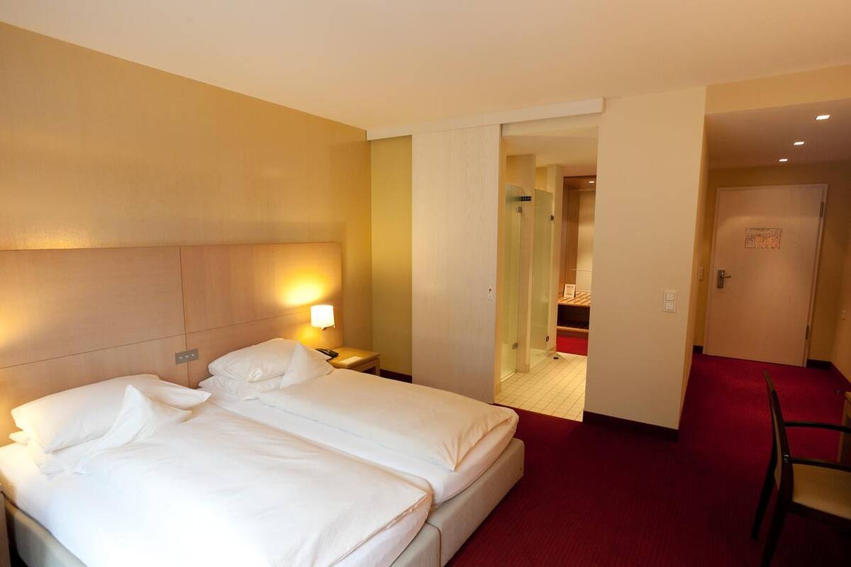 Ein Hotelzimmer mit zwei Betten, einem roten Teppich, einem Holz-Kopfteil, einem Nachttisch mit Lampe und einer Tür zu einem Badezimmer.