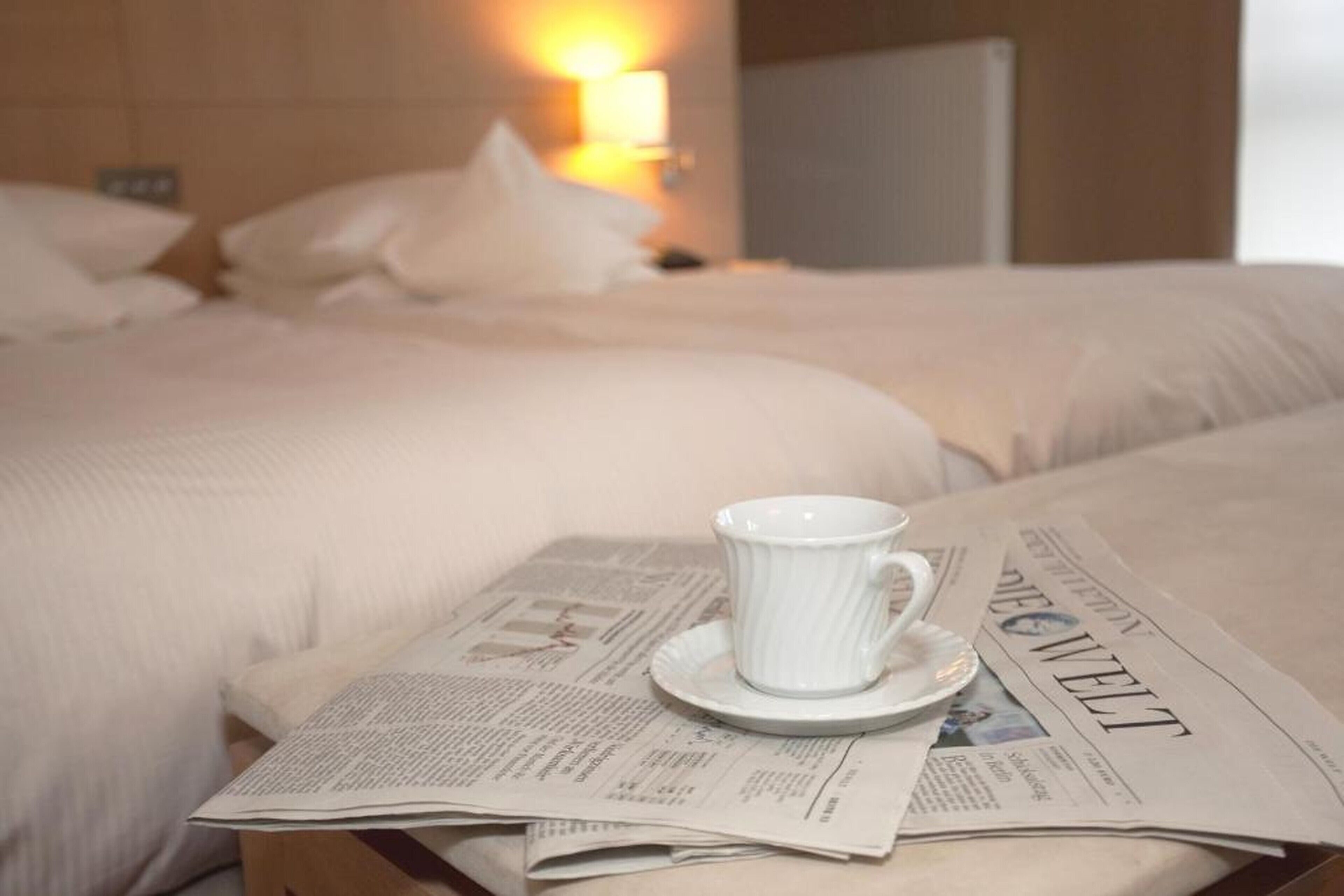 A hotel room with a bed, a cup on a saucer, and a newspaper.